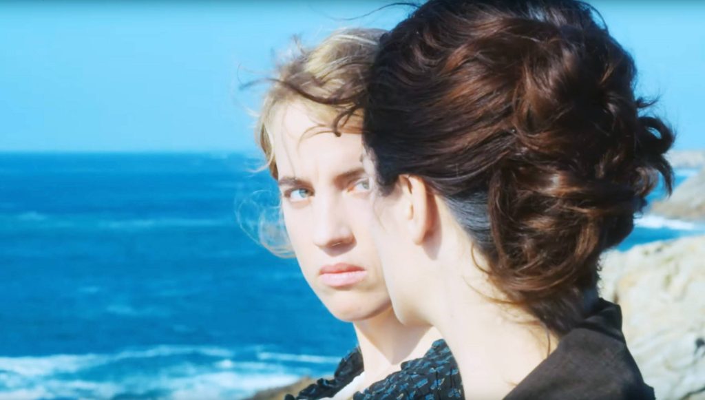 Film still from Portrait of a Lady on Fire. Blonde character looking at brunette, standing near the ocean and cliffs.